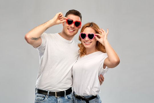 summer accessory, valentines day and love concept - portrait of happy couple in white t-shirts and heart shaped sunglasses over grey background