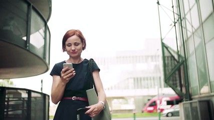 Wall Mural - Young woman commuter with smartphone standing outdoors in city.