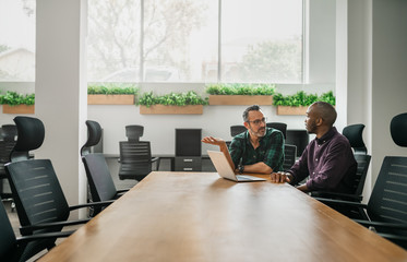 Two businessmen sitting at boardroom table for meeting discussing information on laptop with copyspace