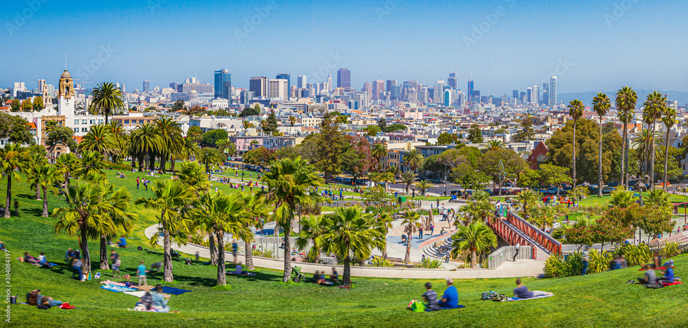 Panoramic view of local people enjoying the sunny summer weather at Mission Dolores Park on a beautiful day with clear blue sky with the skyline of San Francisco in the background, California, USA