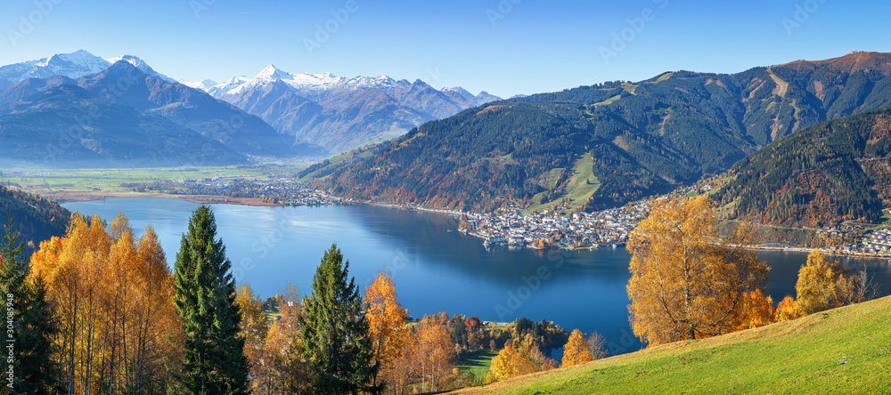 Panoramic view of beautiful autumn scene in the Alps with crystal clear Zeller lake, golden leaves and famous Kitzsteinhorn on a sunny day with blue sky in fall, Zell am See, Salzburger Land, Austria - obrazy, fototapety, plakaty 