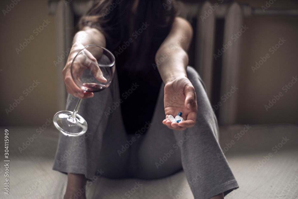a young girl sitting in a dirty room, with a glass of wine, and holds pills in her hand. Poisoning when mixing alcohol and drugs. Polydrug addiction in women