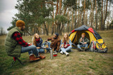 Fototapeta  - Group of friends on a camping or hiking trip in autumn day. Men and women with touristic backpacks having break in the forest, talking, laughting. Leisure activity, friendship, weekend. Eat and drink.