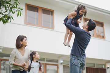 Young Asian family love, father mother and daughter standing playing and fun at outdoor in front of home. father lift up daughter which smiling and felling happy. background is white house.