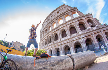 Wall Mural - Happy young man tourist with bike wearing shirt and hat standing on a column taking pictures at colosseum in Rome, Italy at sunrise.