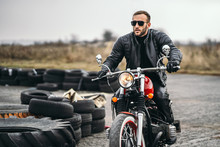 Bearded Man In Sunglasses And Leather Jacket Sitting On A Red Motorcycle And Looking On The Road. Sideways Along The Road Lie Tires