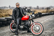Red motorbike with rider. A man in a black leather jacket and pants stands sideways in the middle of the road. Tires are laid on the background