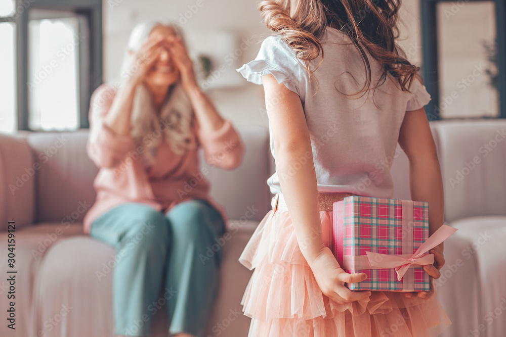 girl holding present box in the hands behind her back and making surprise for grandmother sitting on the couch - obrazy, fototapety, plakaty 