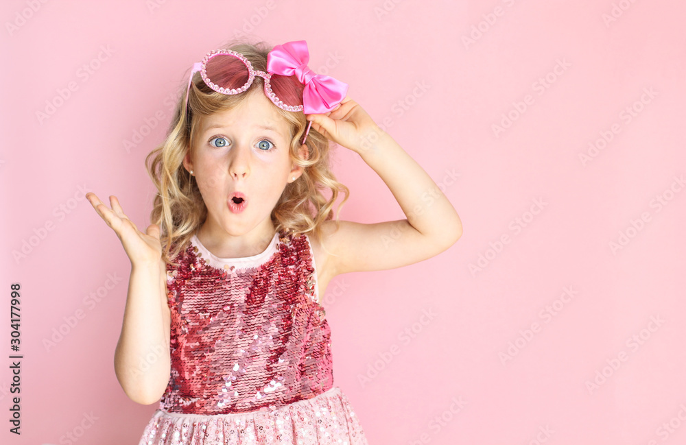 Portrait of a shocked girl wearing pink shiny dress and glasses. Little girl making a surprised expression. Bright and festive background. Party concept. - obrazy, fototapety, plakaty 