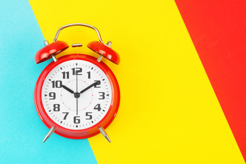 Red retro alarm clock with big dial, on divided diagonally blue-yellow-red background. The concept of time, delay, morning rise, the appointed meeting. Layout with copy space for your text. Flat lay.