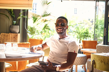 Wall Mural - Happy smiling man in sunglasses sitting at cafe while chatting by mobile phone.