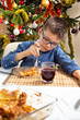 Chłopiec w okularach siedzi przy stole i nabija na widelec jedzenie leżące na talerzu. Pięknie udekorowana choinka w tle.