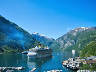 Spectacular view of Geiranger Fjord and mountains with snow, waterfalls, moored cruise ships and tender boats carrying tourists from ship to shore on a beautiful day with blue sky.