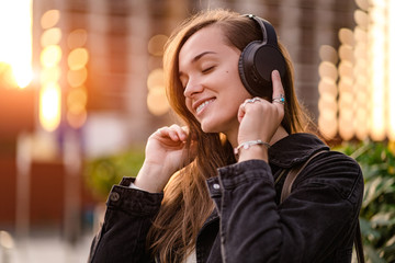 Wall Mural - Happy joyful stylish casual fashionable hipster student woman teenager enjoys listening music using black wireless headphones while walking around the city. Music lover