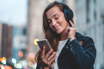 Wall Mural - Young happy stylish trendy casual hipster woman changes songs and tracks on smartphone during listening to music on a wireless headphone while walking around the city. Music lover enjoying music