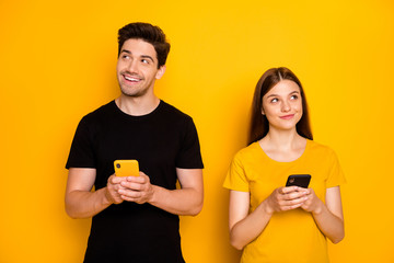 Photo of two fun positive cute nice husband wife people together stylish trendy pondering over what to type holding phones browsing wearing black t-shirt isolated yellow vivid color background