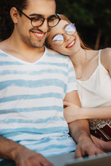 Wall Mural - Lovely young female with red hair looking at camera laughing while embracing his boyfriend and leaning head on his shoulder while he is working at his laptop.