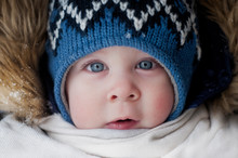 Face Of A Beautiful Baby With Blue Eyes Close-up, In A Blue Knitted Hat, In A White Scarf And A Hood With Fur, Frozen Pink Cheeks In Winter