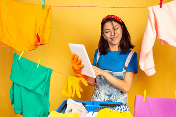 Funny and beautiful housewife doing housework isolated on yellow background. Young caucasian woman surrounded by washed clothes. Domestic life, bright artwork, housekeeping concept. Using tablet.