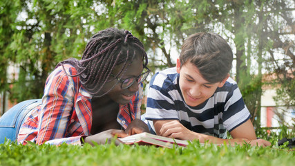 Wall Mural - African teenager girl lying on the grass with her caucasian friend making school lesson. Happyness and Lightheartedness concept