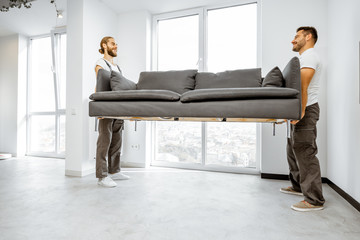 Two professional movers in workwear placing large couch in the living room of the modern white apartment. Furniture delivery concept