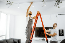 Two Repairmen Or Professional Electricians Installing Light Spots, Standing On The Ladder In The Living Room Of The Modern Apartment