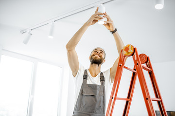 Repairman or professional electrician in workwear installing light spots, standing on the ladder in the white living room