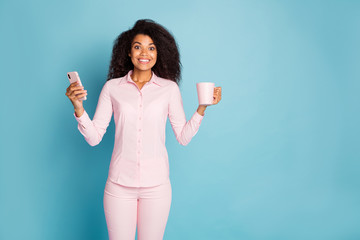 Poster - Photo of pretty wavy dark skin lady holding telephone and hot beverage mug enjoy amazing morning wear pink shirt trousers isolated blue color background