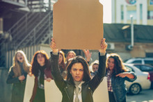 Young Woman With Blank Poster In Front Of People Protesting About Women's Rights And Equality On The Street. Meeting About Problem In Workplace, Male Pressure, Domestic Abuse, Harassment. Copyspace.