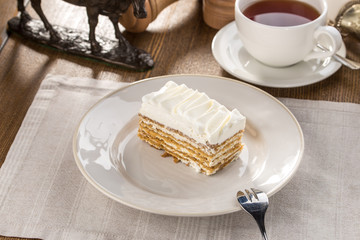 Wall Mural - Slice of layered honey cake with whipped cream and cup of tea on wooden table