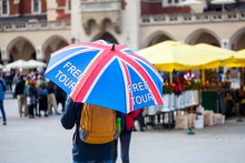 Free Tour On A Cracow Market Square, A UK English Guide With A United Kingdom Flag Umbrella Seen From The Back. Local Free Of Charge Tourist Guides. Tourism, Travelling For England Residents Concept