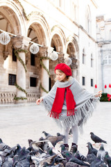 Wall Mural - Cute child girl is feeding doves on european square. Pigeons are flying around little lady. Christmas romantic vintage atmosphere in old town. Authentic, ancient architecture in Dubrovnik, Croatia