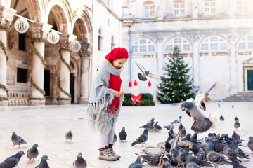 Wall Mural - Kid girl is feeding doves on square with Christmas tree in Europe. Pigeons around cute child. Romantic vintage atmosphere on market in old town. Authentic, ancient architecture in Dubrovnik, Croatia