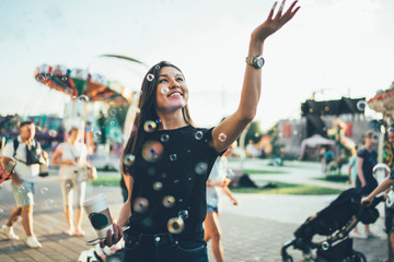 Smiling female with coffee cup visiting public Amusement Park during summer vacations for fall in childhood mood, carefree Asian hipster girl 20 years old touching flying bubbles rejoicing outdoors