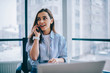 Cheerful young woman in casual wear having telephone conversation sitting at working place near laptop in office, smiling female freelancer calling to colleague for share good news about business.