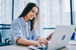 © BullRun - Smiling young female freelancer typing on laptop computer while reading income message on smartphone, positive woman checking notification on mobile phone keyboarding on netbook at desktop in office.