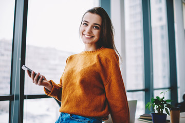 Wall Mural - half length portrait of cheerful female blogger smiling at camera while holding smartphone device du