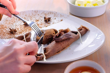 Woman, is eating chocolate russian pancakes, blini with curd filling on the plate. Hands close-up. Served with tea. Close-up hands, side view