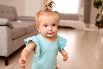 Wall Mural - healthy baby girl in a blue bodysuit in a room next to a gray sofa is learning to walk. the comfort of the home. the child smiles