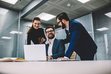 Two businessman and businesswoman take coffee break after conference, team watching funny videos and laughing in their office, colleagues smiling and discussing successful meeting projects together