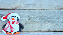 Penguin With Santa Claus Hat, Striped Red And White Scarf, And Orange Feet Laying Flat On Natural Wood Background With Copy Space