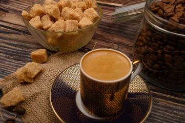 A Cup of coffee, pieces of brown sugar in a sugar bowl, coffee beans in a glass jar on a wooden background.