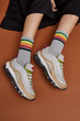 Cropped shot of a girl's foots in white sneakers, lying on a brown background. It is grey artwork socks with colored stripes on foots. 