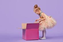 A Funny Little Girl Child In A Magnificent Holiday Dress Look Inside A Large Gift Colored Box, On A Purple Isolated Background. Birthday Surprise