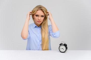 Concept cute model misses losing time sitting at the table. Close-up portrait of a beautiful blonde girl with excellent makeup with long smooth hair on a white background in a blue shirt.