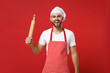Laughing young bearded male chef cook or baker man in striped apron white t-shirt toque chefs hat posing isolated on red background. Cooking food concept. Mock up copy space. Hold wooden rolling pin.