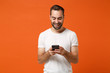 Smiling young man in casual white t-shirt posing isolated on orange background, studio portrait. People sincere emotions lifestyle concept. Mock up copy space. Using mobile phone, typing sms message.