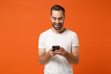 Smiling Young Man In Casual White T-shirt Posing Isolated On Orange Background, Studio Portrait. People Sincere Emotions Lifestyle Concept. Mock Up Copy Space. Using Mobile Phone, Typing Sms Message.