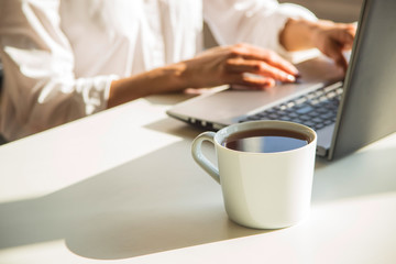 A modern freelancer workplace with a laptop and a cup of tea on a white table in the shadows in a light interior.