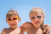 Two Little Girls In Sunglasses Are Having Fun. Girlfriends Hug On The Beach. Portrait Of Two Cheerful Children. Concept Of Travel And Summer Vacation